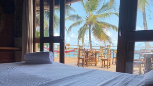 A room with a bed, a window overlooking a beach with palm trees and wooden furniture.