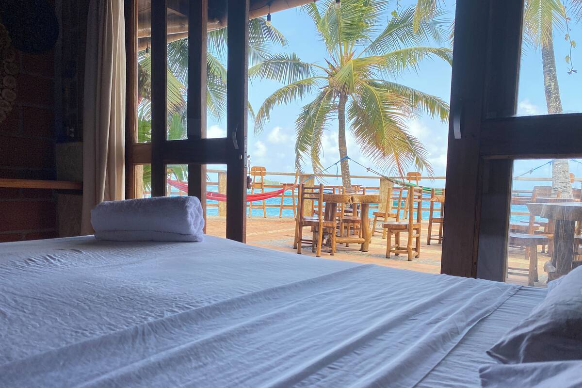 A room with a bed, a window overlooking a beach with palm trees and wooden furniture.