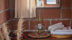 A rustic room with a brick wall, a window with a curtain, a woven basket, a vase with dried plants, a tray with a bottle, glasses, and towels.