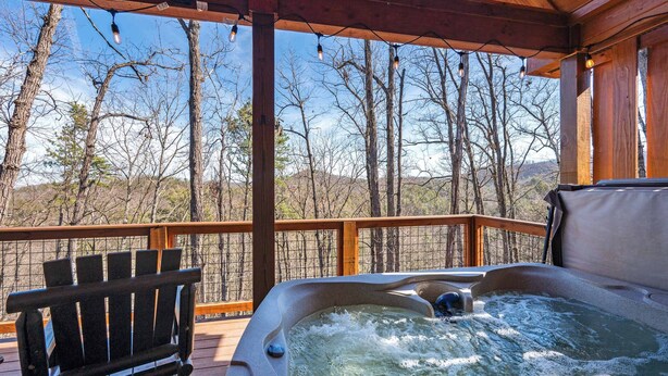 Een hottub op een houten terras met een bankje, omringd door bomen en een heldere lucht.