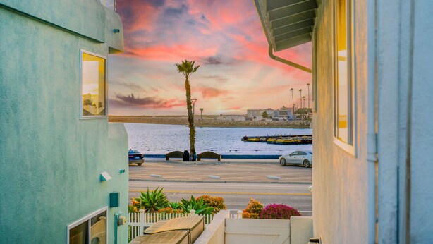 A view from a balcony overlooking a parking area with a palm tree, a road, and a body of water. - Walk To Beach, Amazing Views, Private Jacuzzi* (Oxnard)