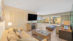 A spacious living room with a large sofa, a coffee table, and a TV. There is a balcony with a view of buildings. - Walk To Beach, Amazing Views, Private Jacuzzi* (Oxnard)