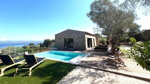 Un espace aménagé au bord de la piscine, avec un bâtiment en pierre, une terrasse en bois et une vue sur la mer.