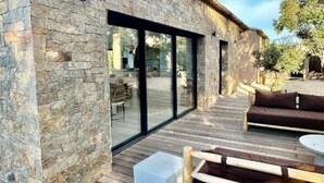 Une façade en pierre, une terrasse en bois, une porte vitrée donnant sur l’intérieur et du mobilier d’extérieur.