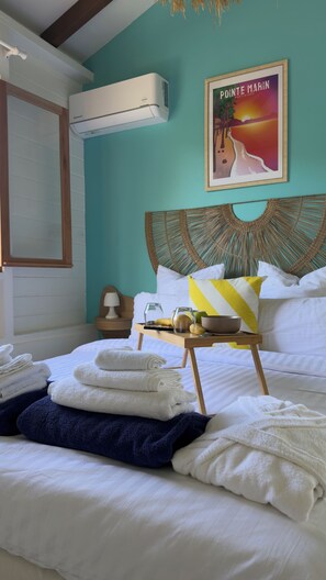 Een netjes opgemaakt bed met witte en marineblauwe handdoeken, een nachtkastje met een lamp en een schaal, en een ingelijst foto van Pointe Marin aan de muur.