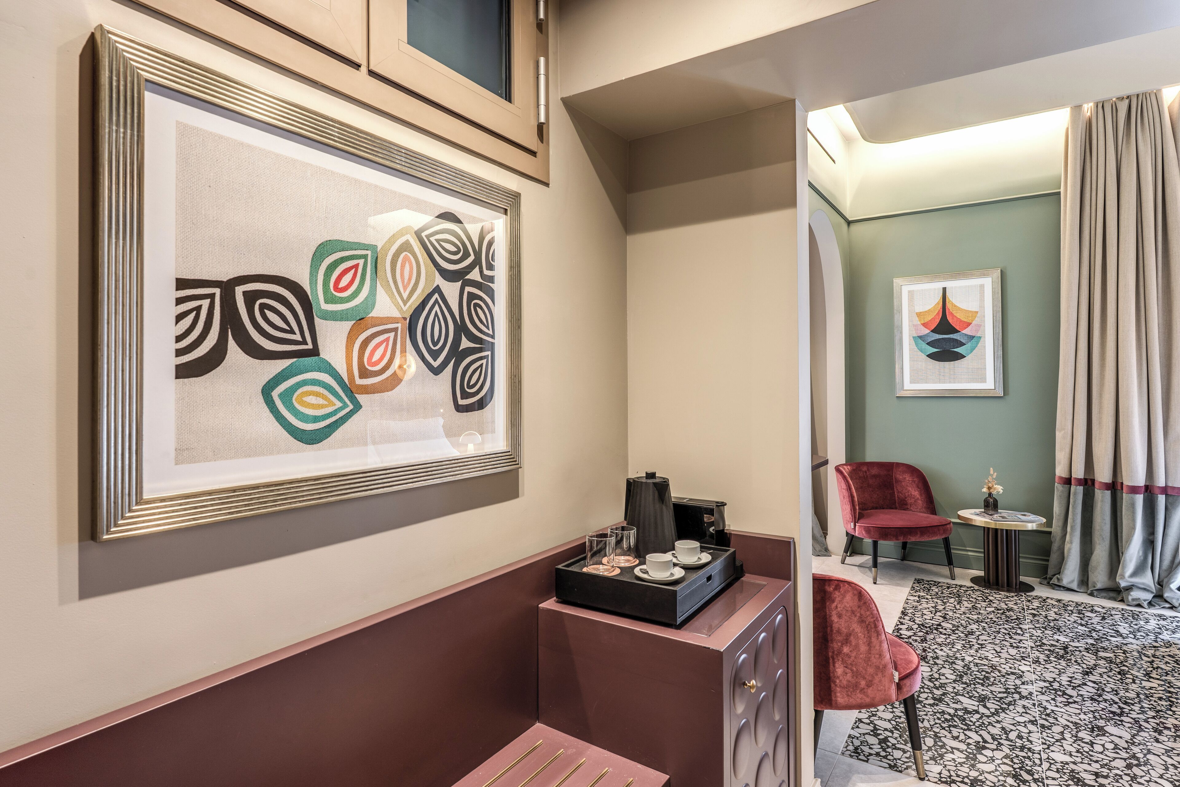 Habitación de hotel con una obra de arte abstracta enmarcada, una mesita con cafetera, una silla roja y una alfombra estampada.