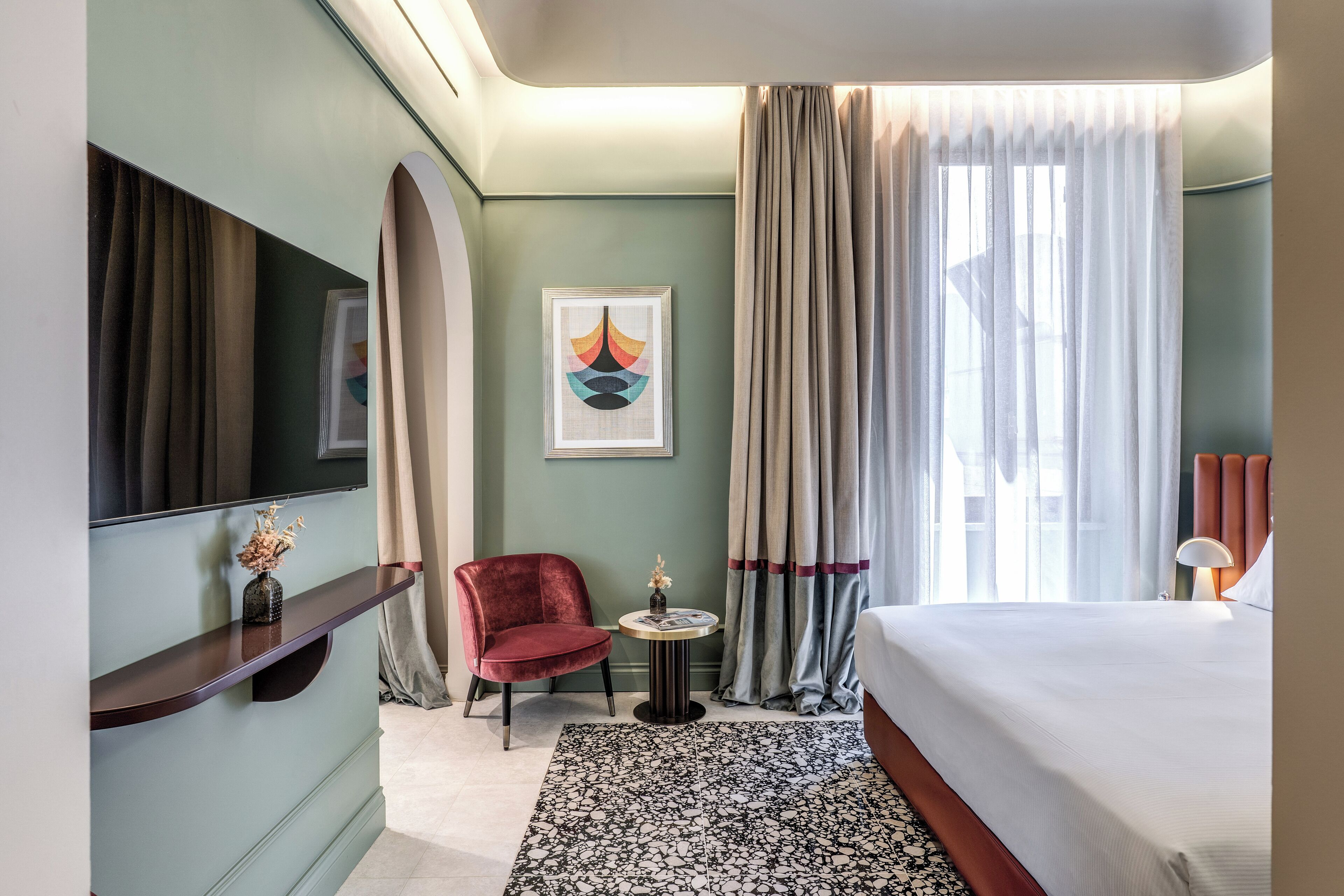 A hotel room with a bed, a TV mounted on the wall, a red chair, a small table with a vase, and a large window with curtains.