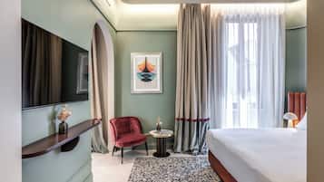 A hotel room with a bed, a TV mounted on the wall, a red chair, a small table with a vase, and a large window with curtains.