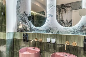 Une salle de bain avec deux lavabos roses, un grand miroir et un mur orné d’une fresque représentant des feuilles tropicales.