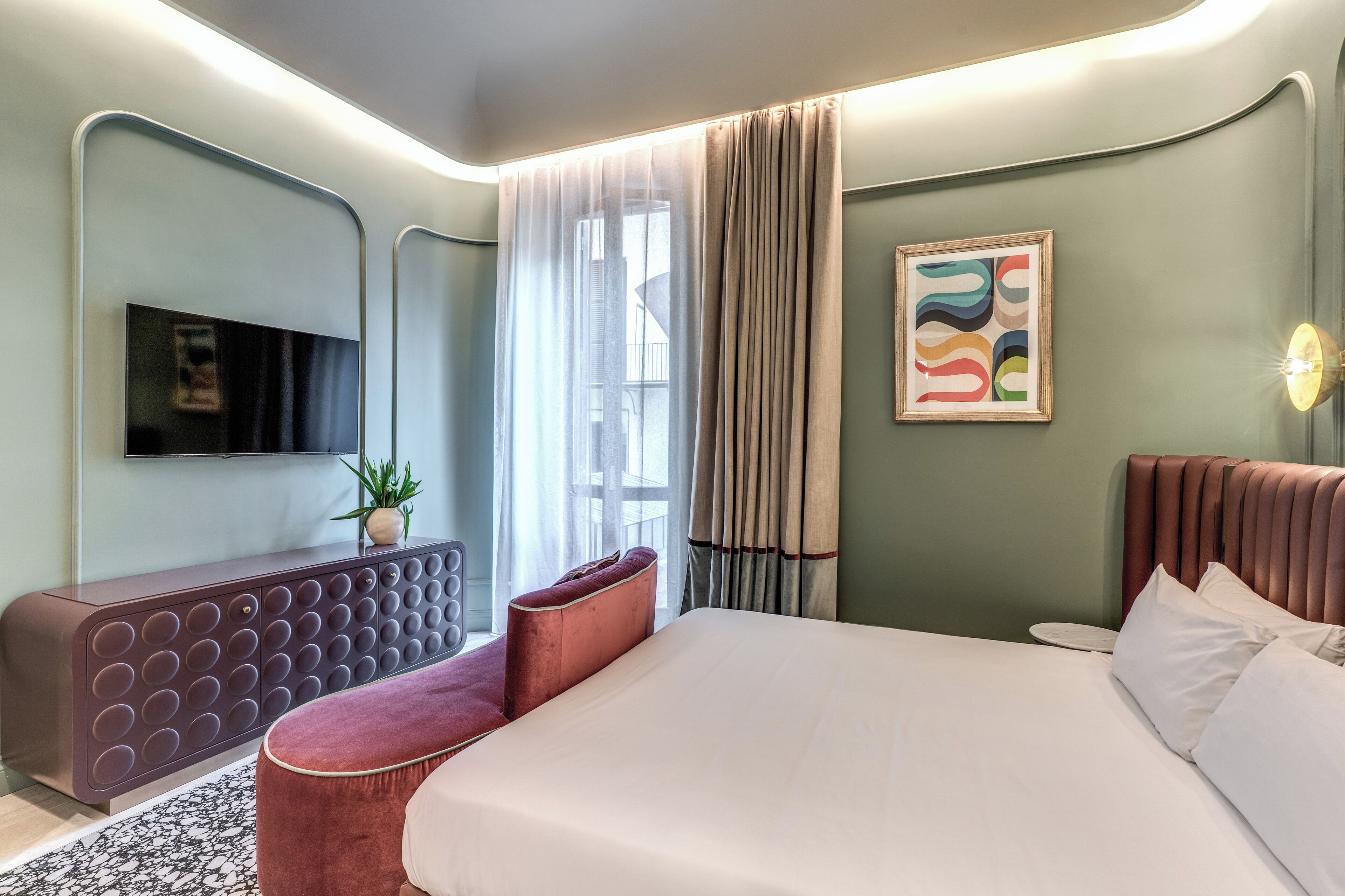 A modern hotel room with a large bed, a seating area with a cushioned bench, a flat-screen TV mounted on the wall, and a potted plant on a console table.