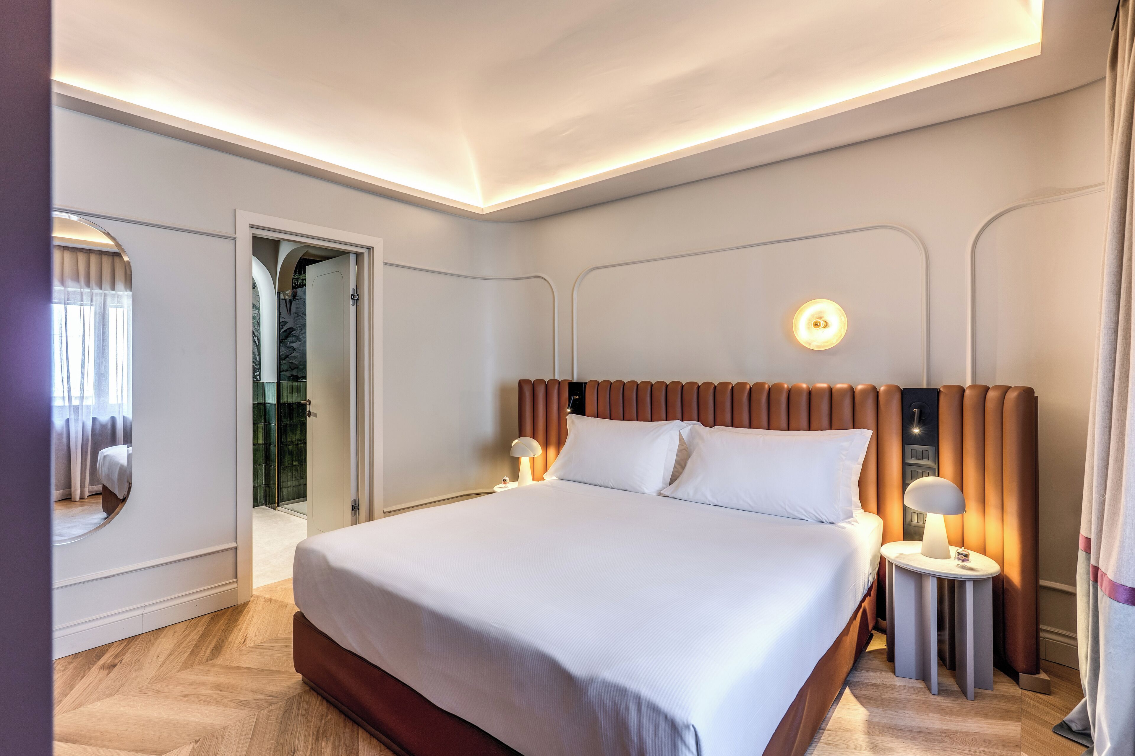 A modern hotel room with a large bed, a bedside table with a lamp, and a view of a bathroom with a shower and glass door.
