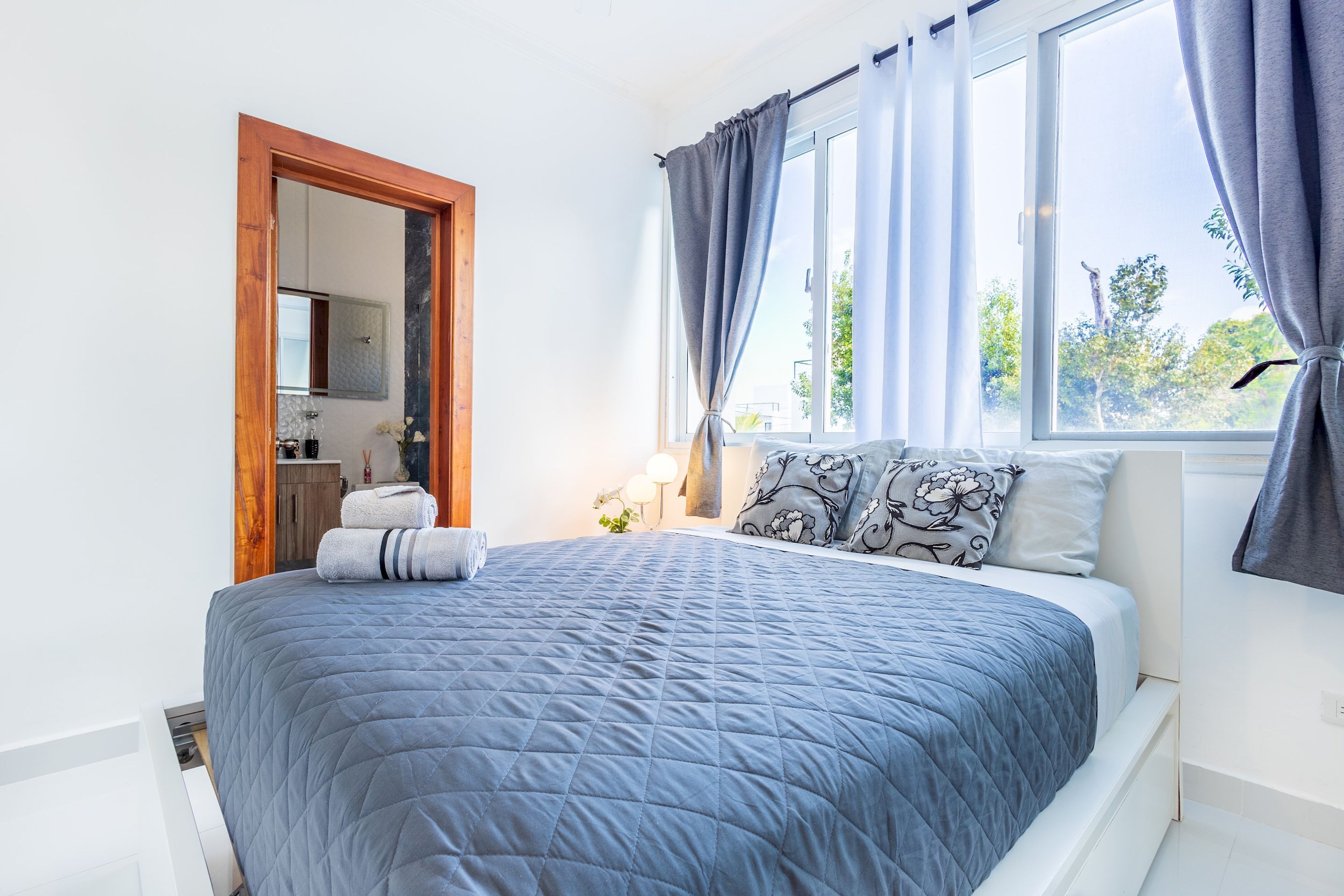A bedroom with a bed, a window with curtains, a nightstand, and a mirror.