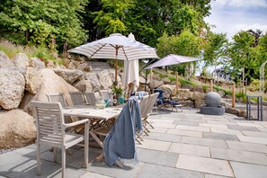 An outdoor patio with stone flooring, wooden chairs, and two umbrellas.