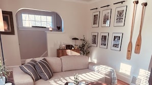 Una sala de estar con un sofá, otomanas, una mesa de centro y una silla de madera. Hay cuadros enmarcados en la pared y una ventana con persianas.