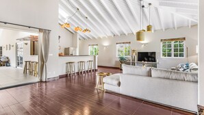 A spacious living room with a high ceiling, wooden flooring, a large sofa, a TV, and a kitchen area with stools. - Detached Villa with private pool within walking distance to the beach & restaurants (Jan Thiel)