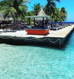 A beachside resort with a thatched-roof gazebo, lounge chairs, and a wooden pier extending into the turquoise sea.