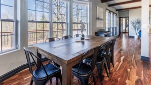 A spacious dining area with a long wooden table and black chairs, large windows with a view of trees, and a piano in the background.