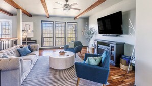 A spacious living room with a fireplace, a television mounted on the wall, and a large window with a view of trees.