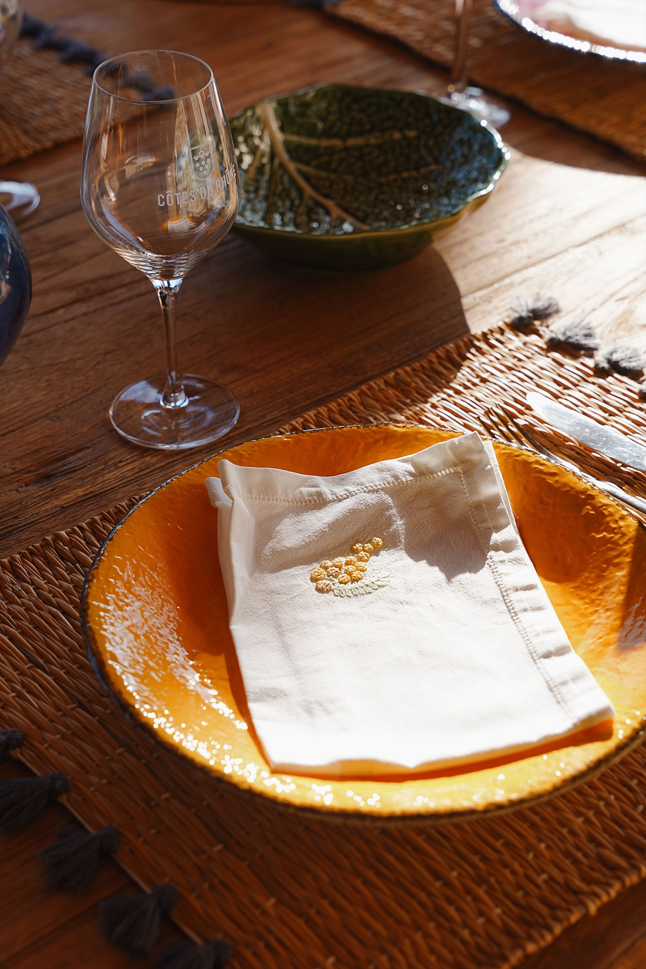 A table with a clear glass wine glass, a green ceramic bowl, and an orange plate with a folded napkin.