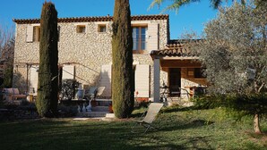 Un bâtiment en pierre avec un toit de tuiles, deux cyprès imposants et une pelouse où se trouve une seule chaise.