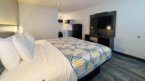A modern hotel room with a large bed, a built-in desk, a flat-screen TV, and a kitchenette.
