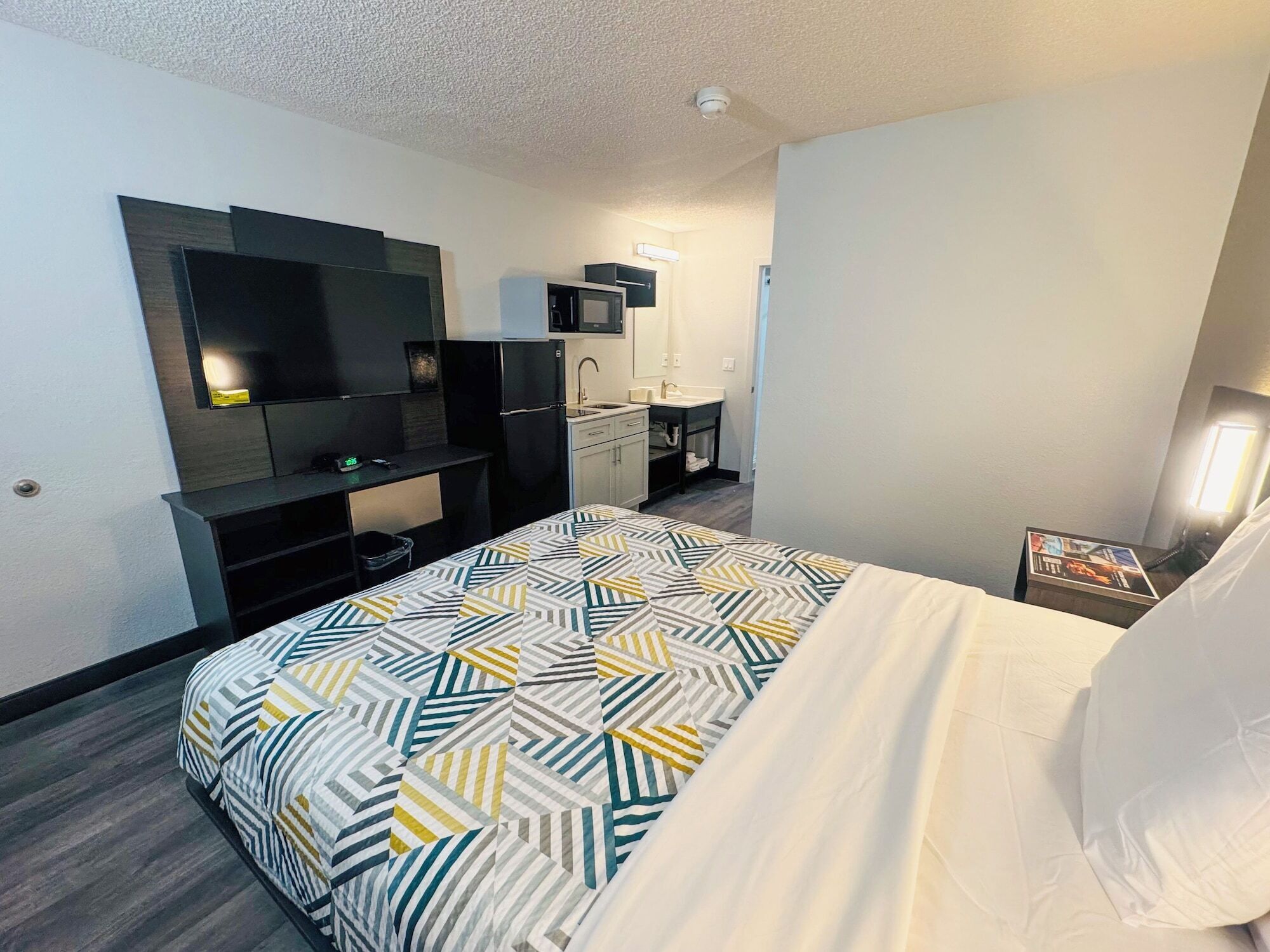 Une chambre d’hôtel équipée d’un lit, d’une télévision, d’un four à micro-ondes, d’un lavabo et d’une petite table avec un magazine.