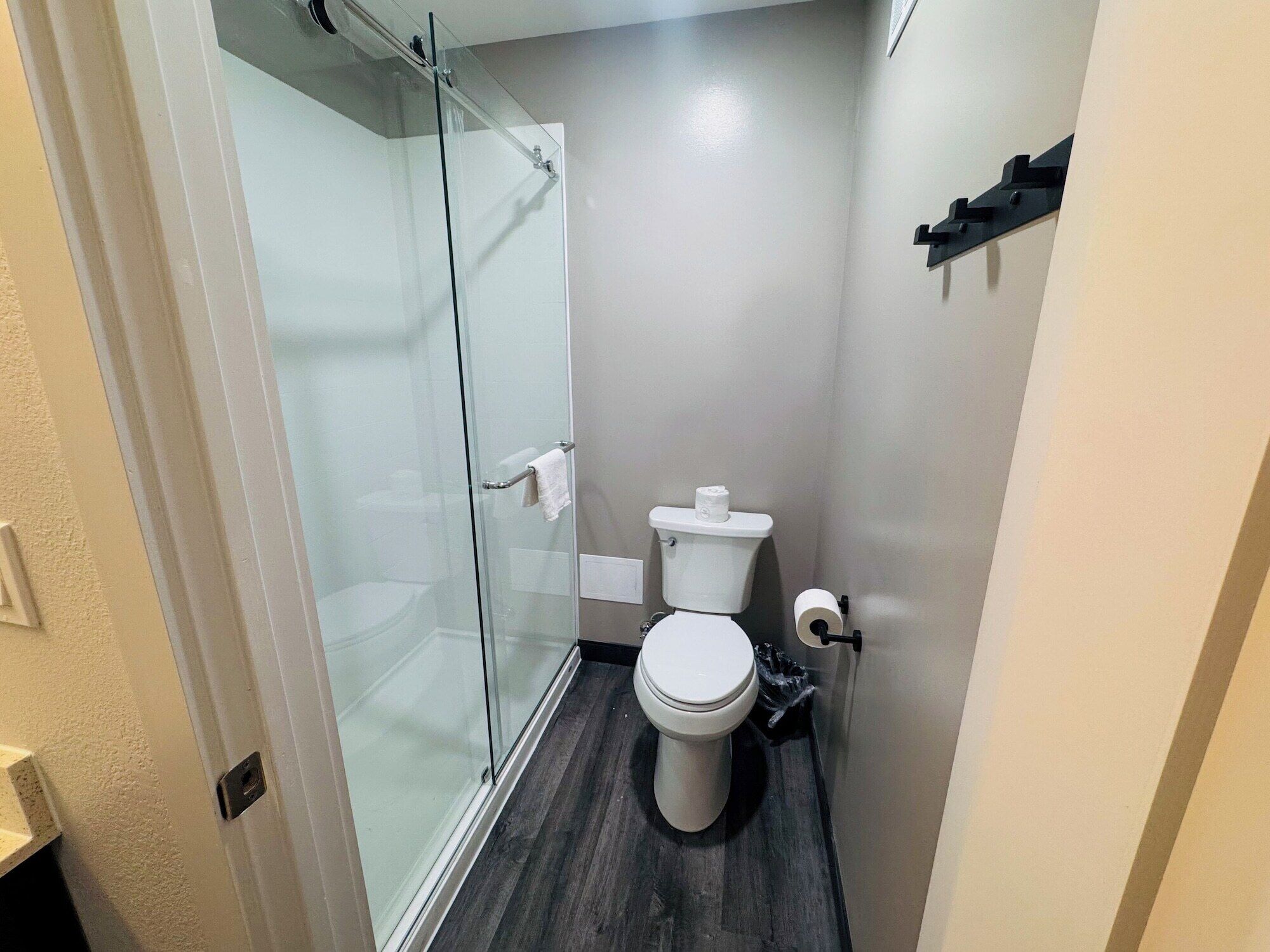 A small, modern bathroom with a glass shower enclosure, a white toilet, and a dark wood floor.