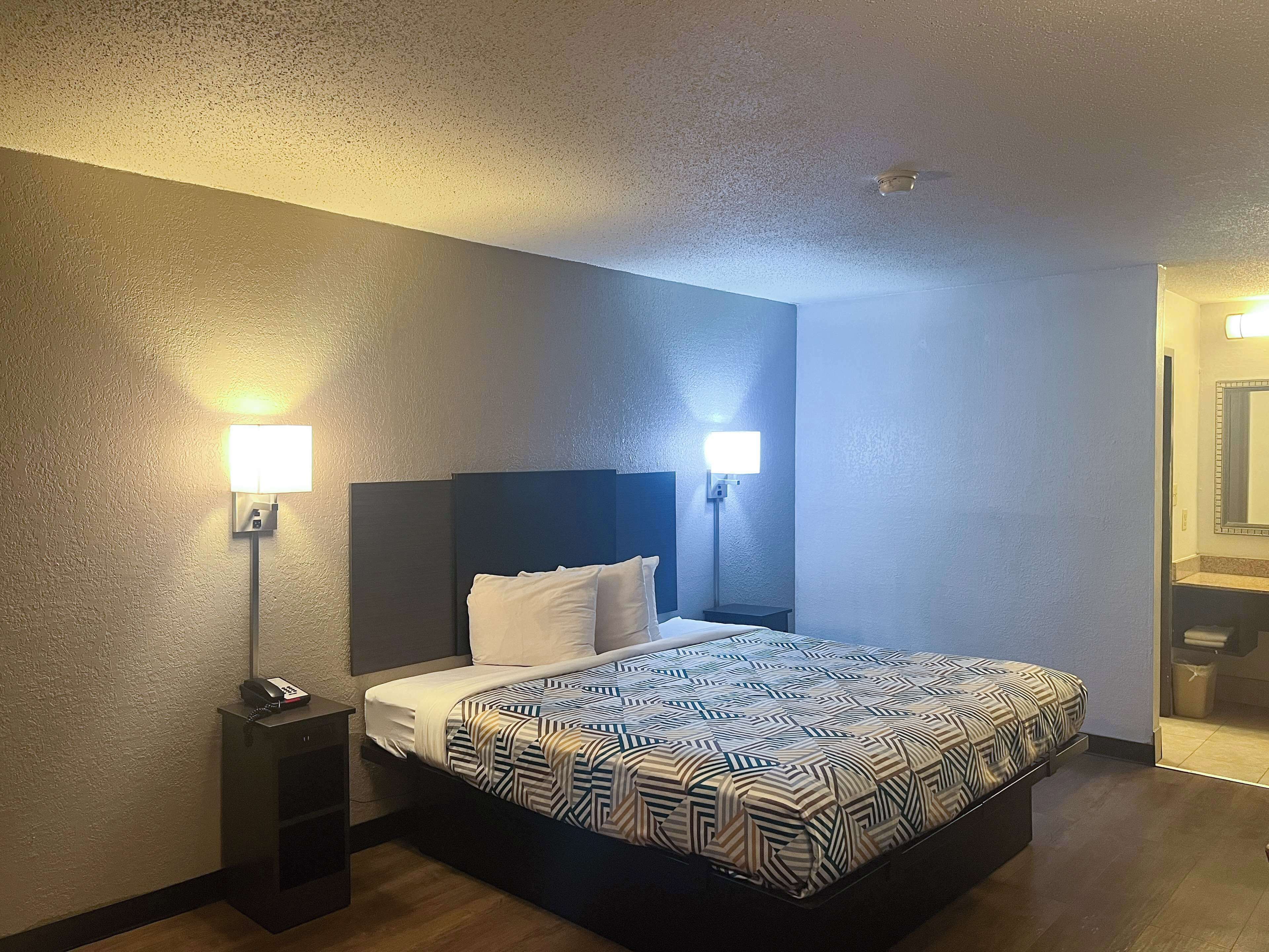 A hotel room with a bed, bedside lamps, a nightstand, and a bathroom visible through an open door.