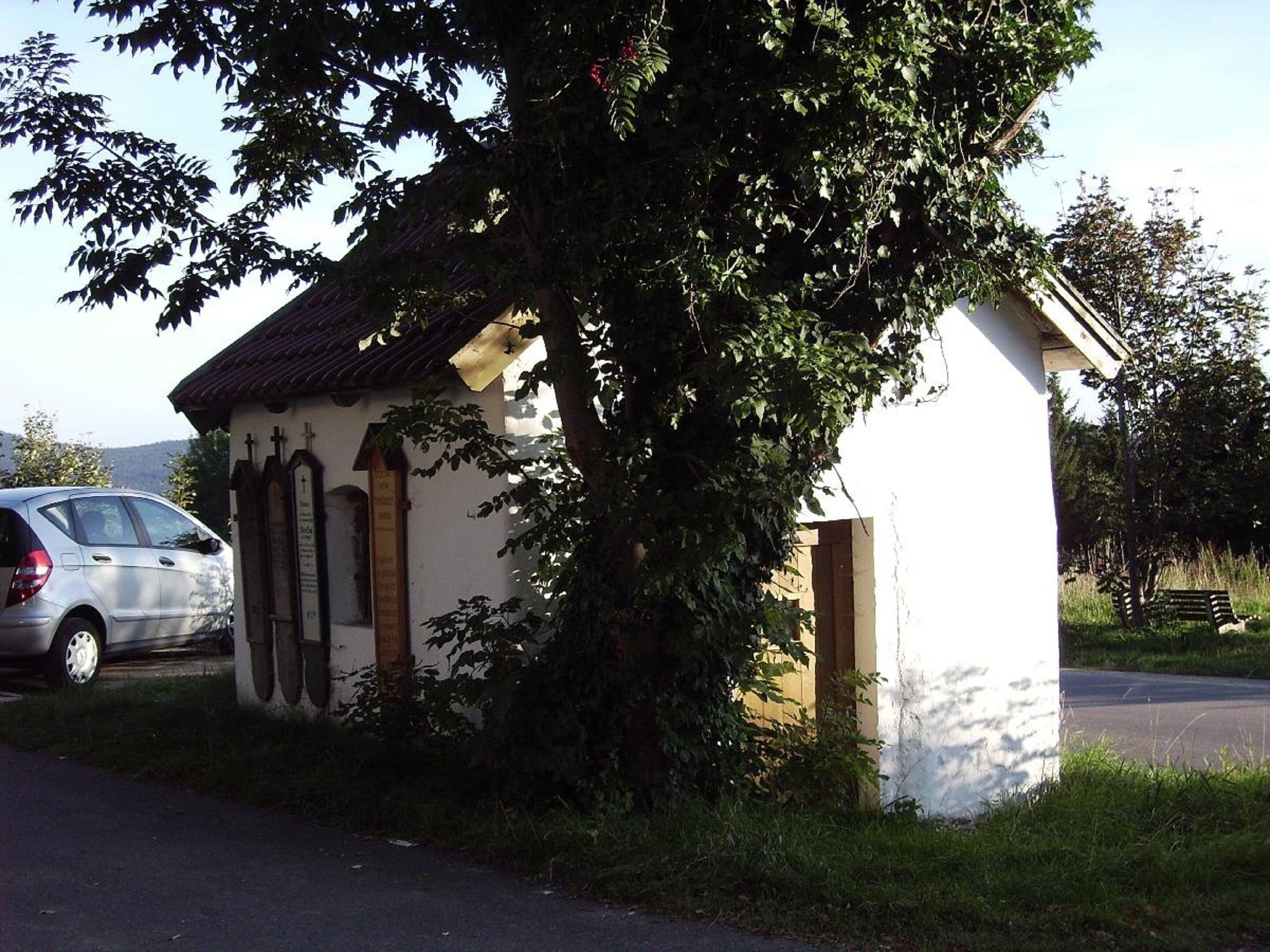 Un petit bâtiment doté d’un toit en pente, qui pourrait être une chapelle ou un centre communautaire, entouré de verdure et avec une voiture garée à proximité.