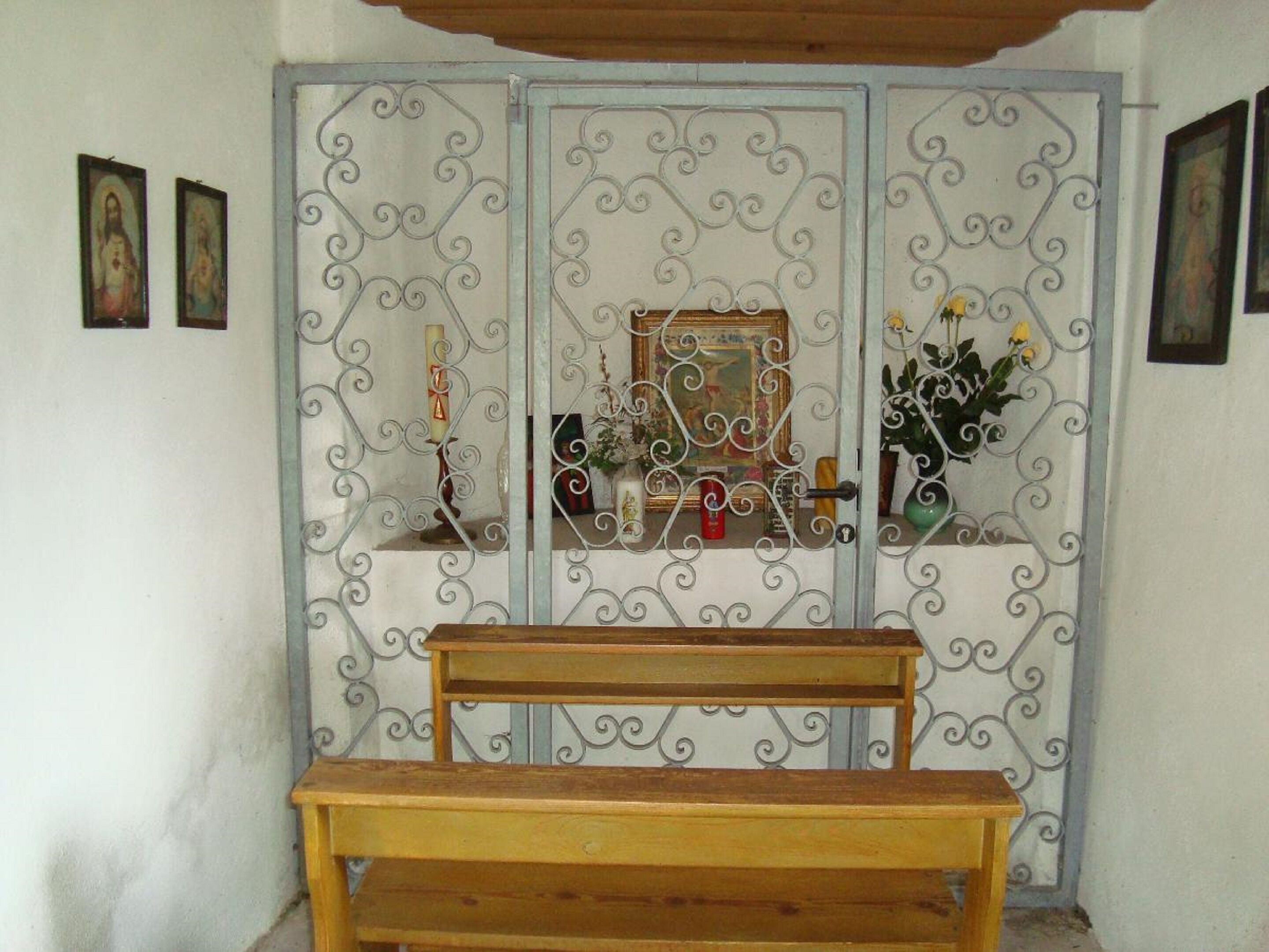 A room with a wooden bench in the foreground, a decorative metal gate with intricate designs, and a small shelf with various items including a candle, a framed picture, and a vase with flowers.