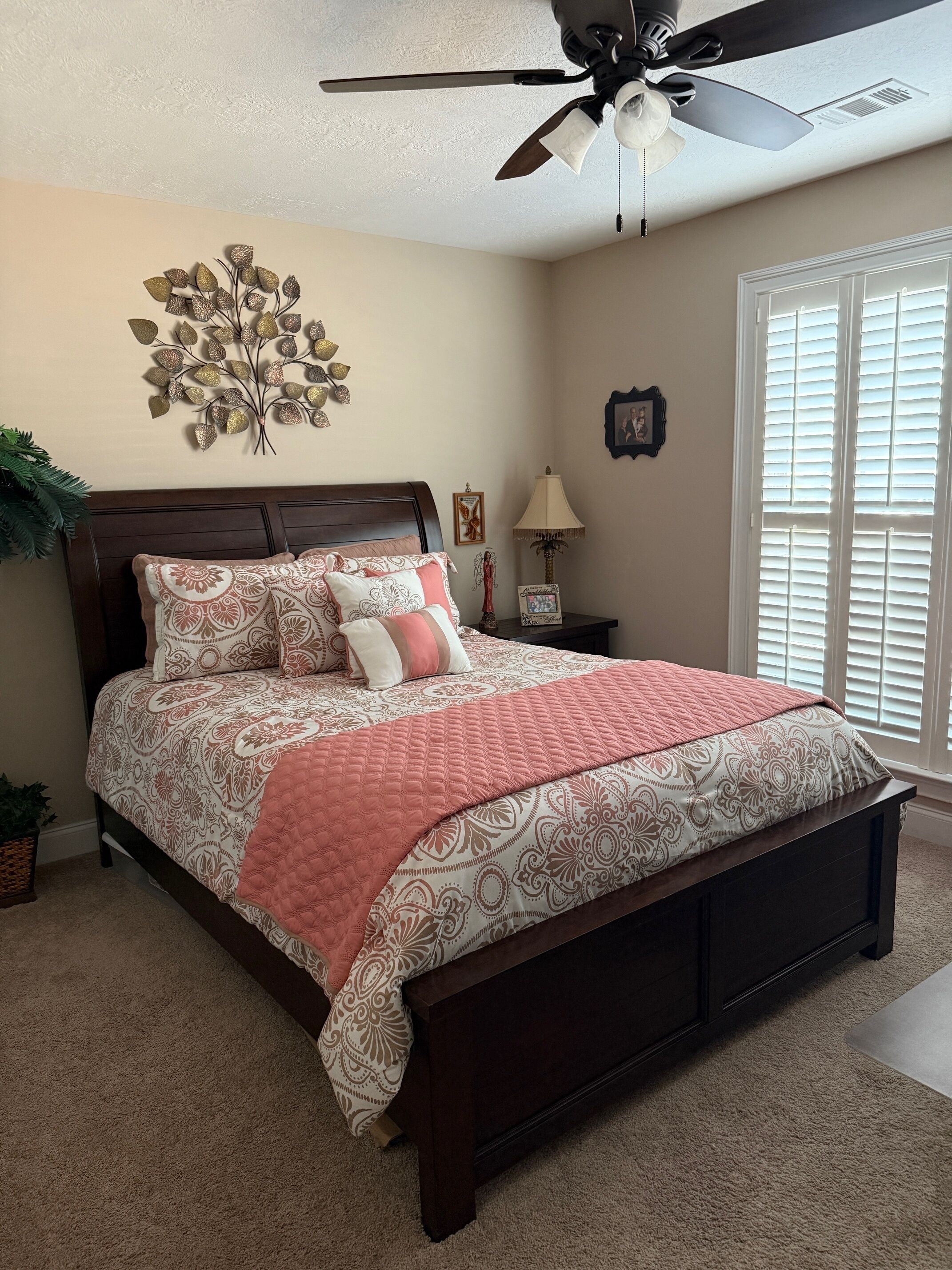 A bedroom with a bed, a ceiling fan, a wall decoration, a nightstand, a lamp, and a window with blinds.