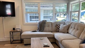 Un salon moderne avec un canapé d’angle, une table basse, un téléviseur à écran plat et une grande fenêtre offrant une vue sur l’extérieur.