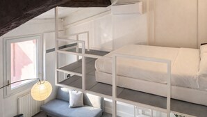 Une chambre à coucher de taille réduite, décorée dans des tons de blanc, avec un lit superposé, un petit coin salon et une fenêtre laissant entrer la lumière naturelle.