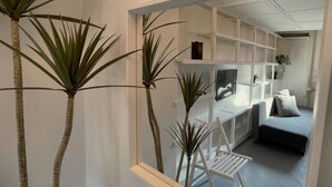 Un intérieur moderne avec un bureau blanc, un lit et une chaise. On aperçoit des plantes au premier plan.