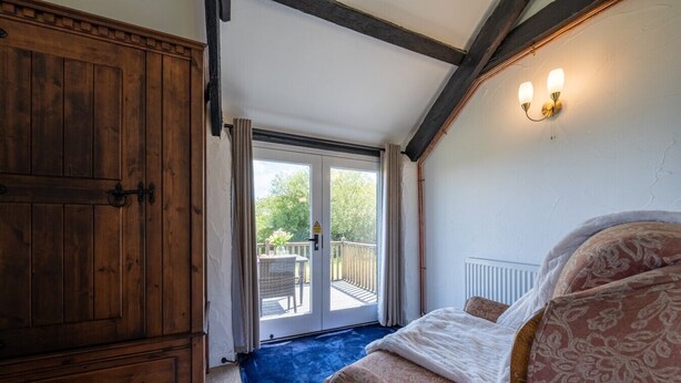 A bedroom with a large wooden wardrobe, a bed with patterned bedding, a blue rug, and a balcony with a table and chairs. - Bligh - cosy cottage near Crackington Haven (Cornwall)