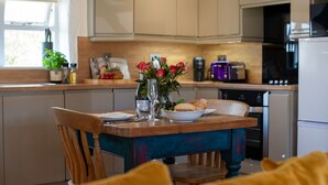 A kitchen with wooden cabinets, a dining table with chairs, a vase with flowers, and a toaster. - Bligh - cosy cottage near Crackington Haven (Cornwall)
