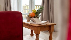 A wooden table with a book, a fruit bowl, and a red chair in a room with a view through a window. - Butterwell - Treworgie Barton Cottages (Cornwall)