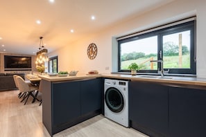 A modern kitchen with a built-in washing machine, a dining area with a sofa, and a large window offering a view of the outdoors. - Manor a spacious modern cottage for families or groups (Cornwall)