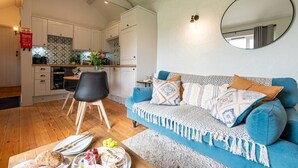 Un salon moderne avec un canapé bleu, une table à manger avec des chaises, une cuisine en arrière-plan et un miroir rond accroché au mur.