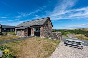 A stone building with a sloped roof, a paved entrance, and a picnic table in the foreground. - Trespaddick - Treworgie Barton Cottages (Cornwall)