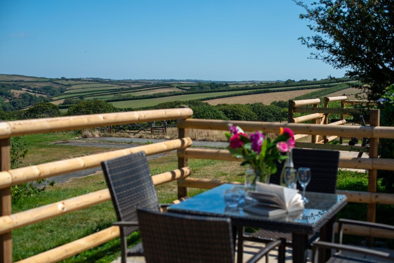 An outdoor dining area with a view of rolling hills, wooden railings, and a table set with a vase of flowers.