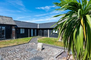 A building with a stone pathway leading to a set of doors, surrounded by grass and a palm plant. - Braddon - Treworgie Barton Cottages (Cornwall)