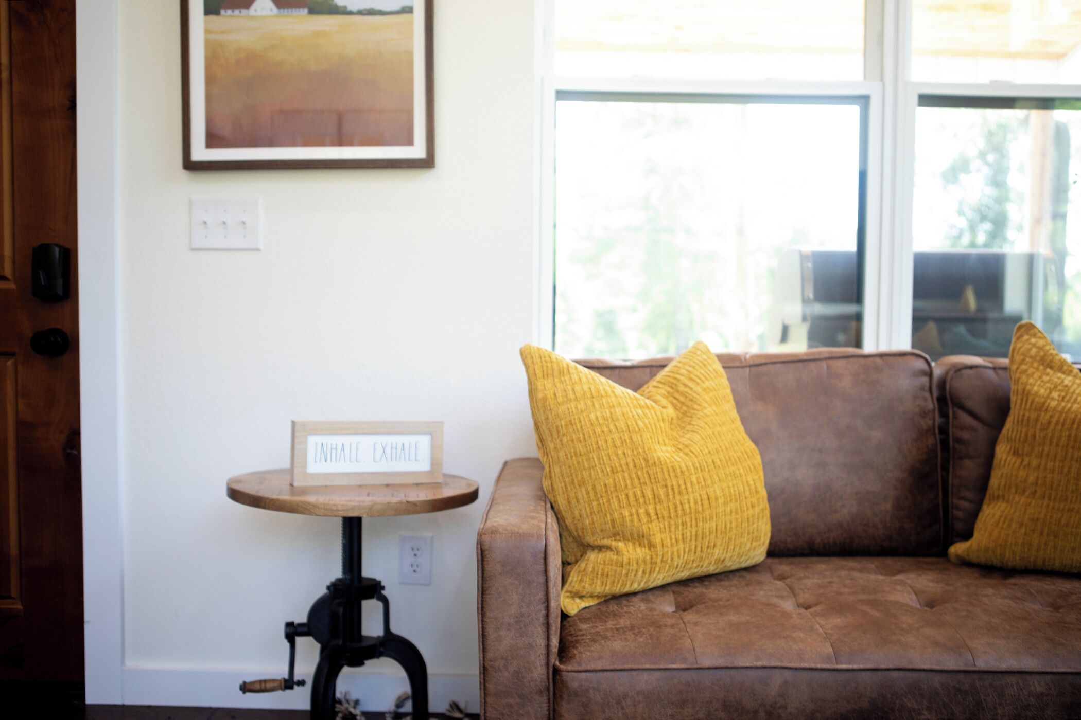 A brown leather sofa with two yellow cushions, a small wooden table with a sign, and a framed picture on the wall.