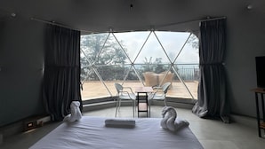 Una habitación de hotel moderna con una cama grande, una mesita y dos sillas. La habitación tiene vista a un balcón con barandilla y árboles.