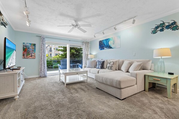A living room with a ceiling fan, a flat-screen TV, a sofa, a coffee table, and a view of a balcony. - Beach View condo w/ private beach access & sunset views (Siesta Key)