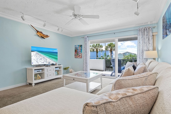 A living room with a television, white furniture, a ceiling fan, and a view of the outdoors.