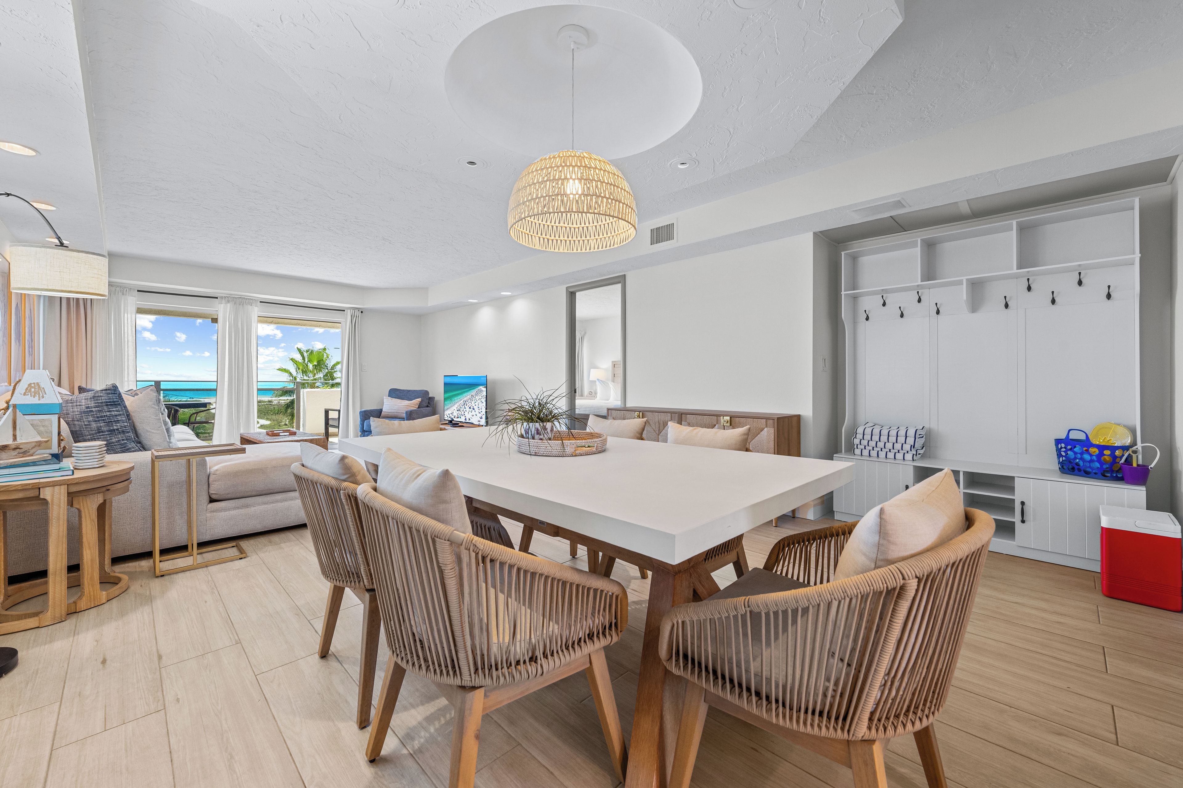 A spacious living room with a large dining table, wicker chairs, a ceiling light, and a view of the ocean.