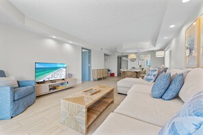 A modern living room with a flat-screen TV, a sofa, a coffee table, and a dining area in the background.