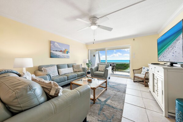 A living room with a sofa, armchair, coffee table, and a television. There is a balcony with a view of the ocean.
