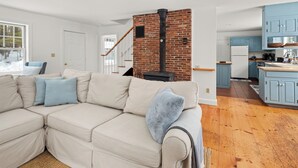 A modern living room with a beige sectional sofa, a brick fireplace, and a kitchen with blue cabinets. - Green Mountain Getaway (Waitsfield)